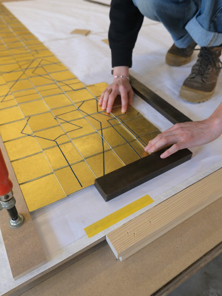 Victoire Camus dans l'atelier de marqueterie de Pierre-Henri Beyssac, pendant le montage de leur cocréation "Memento", marqueterie de papier suspendue, dorée à la feuille d'or